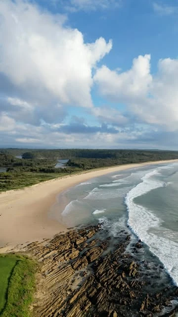 Les vagues longent une grande plage et une avancée rocheuse sous les nuages.