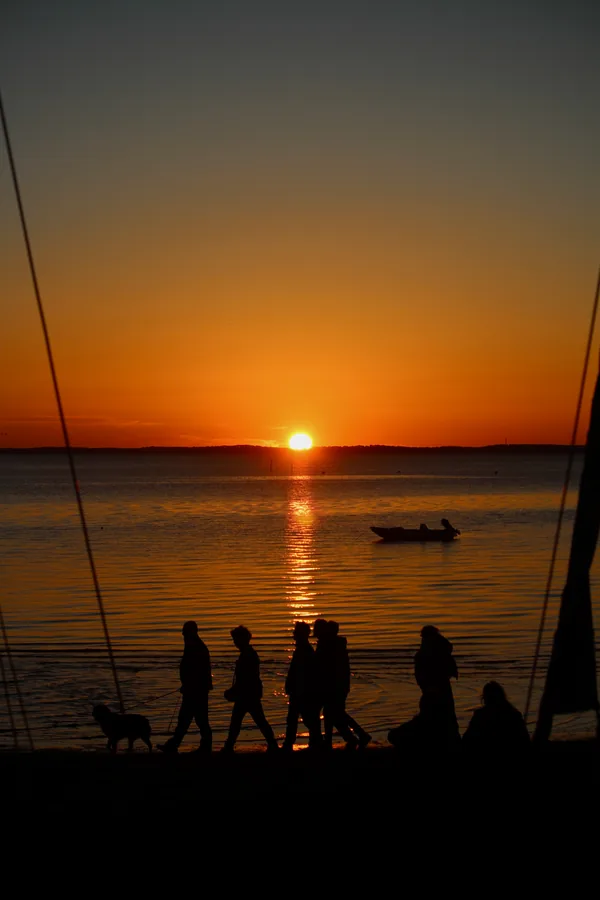 Des silhouettes se promènent au bord de l’eau au coucher du soleil.