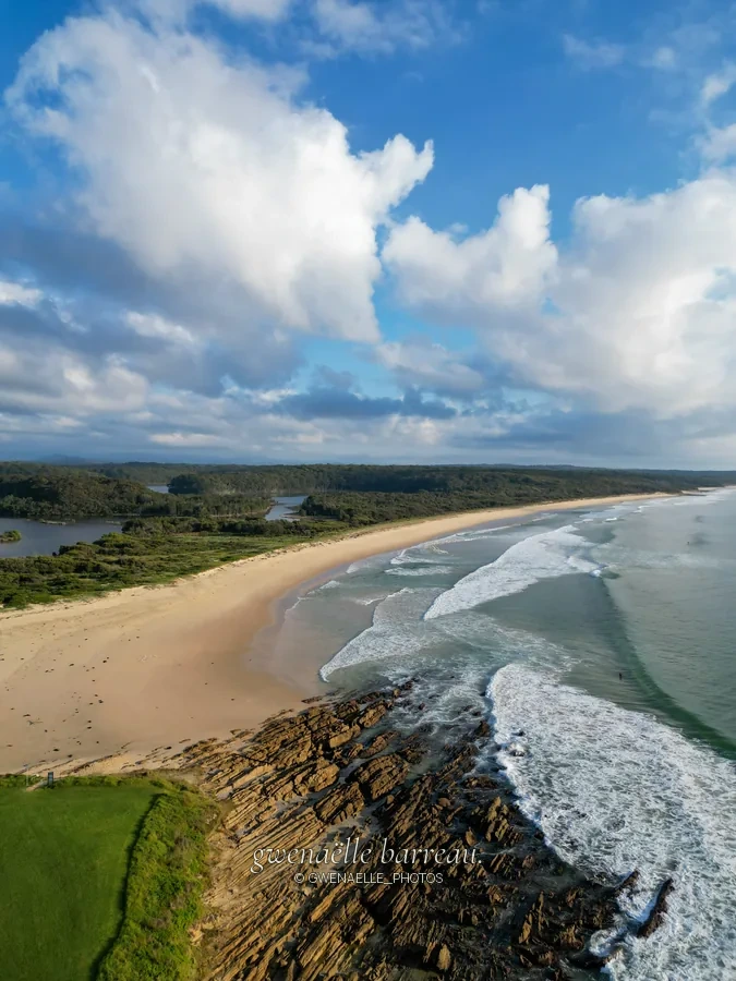 Grande plage incurvée et rochers au premier plan sous des nuages blancs.