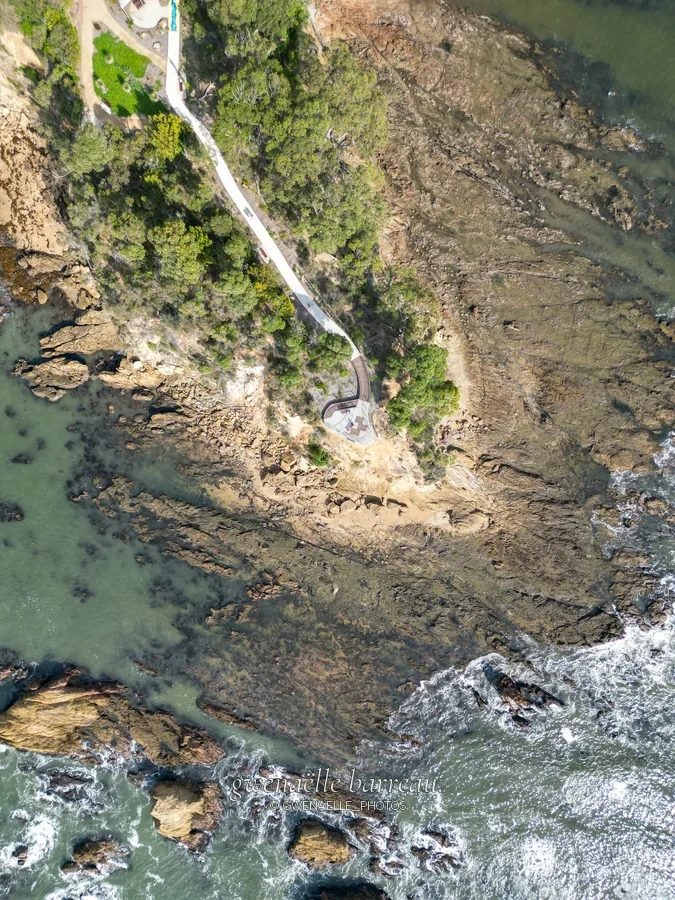 Passerelle côtière et rochers baignés par une eau peu profonde, vus du ciel.