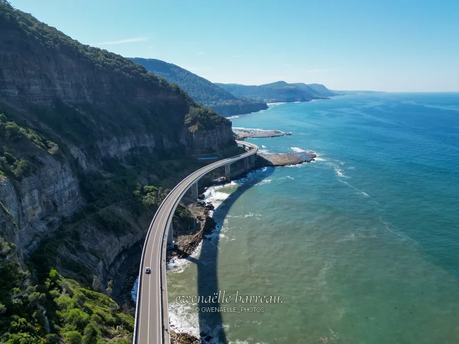 Route courbe longeant une haute falaise au-dessus de la mer bleue.