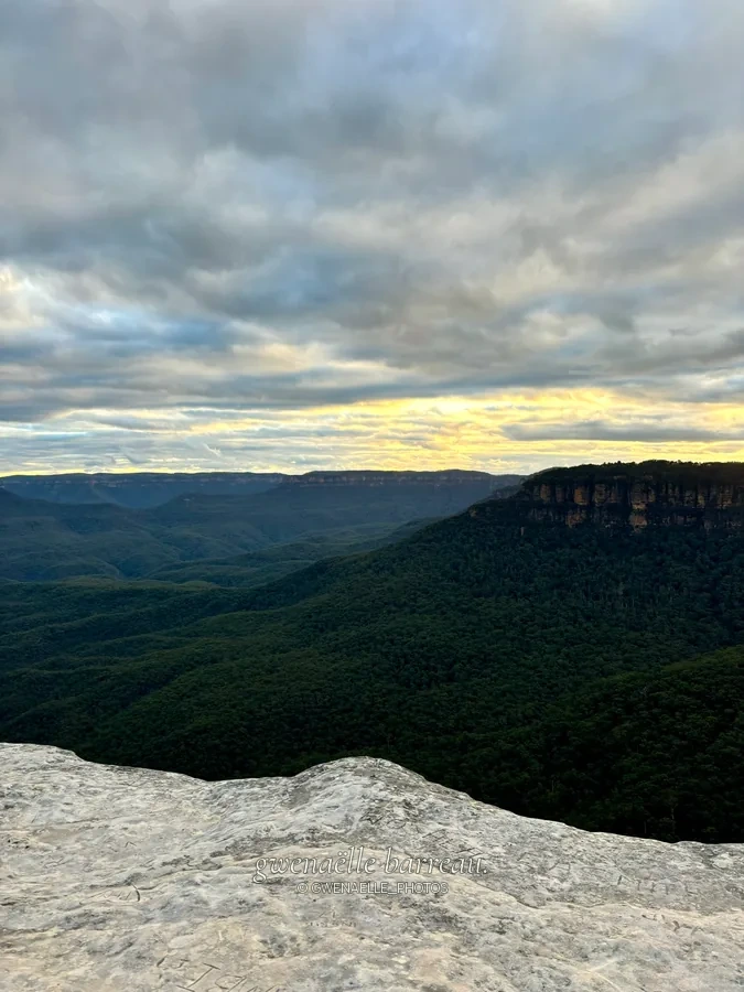 Vallée et plateaux boisés sous un ciel couvert illuminé à l'horizon.