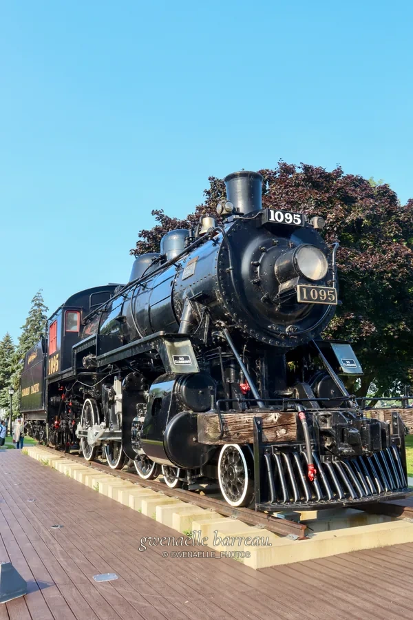 Locomotive à vapeur noire exposée sur des rails sous un ciel bleu.