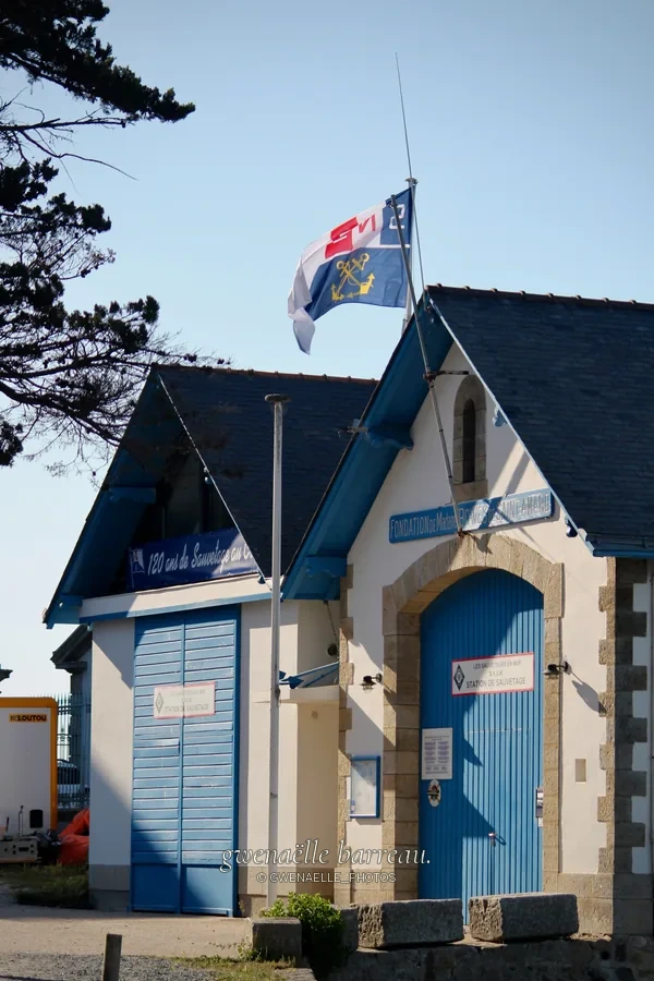 Petite construction aux portes bleues et au toit d'ardoise sous un drapeau.