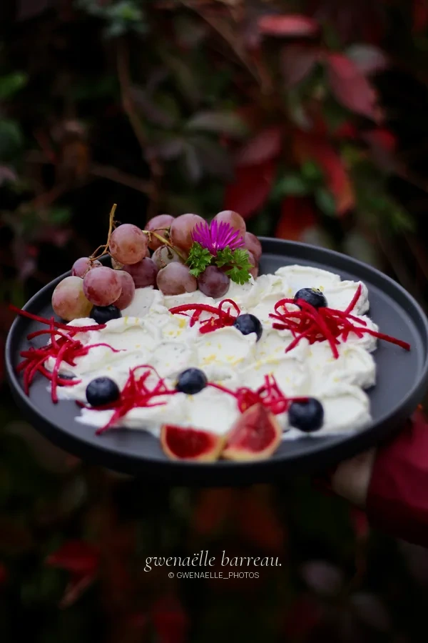 Assiette de fromage frais, fruits et garnitures rouges devant un feuillage sombre.