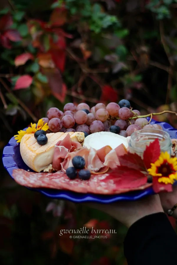 Planche garnie de raisins, fromages, charcuterie et fleurs devant un feuillage sombre.